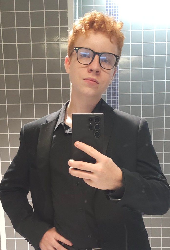 Eli Searle taking a mirror selfie in a black suit — grey tiled fitting room or bathroom