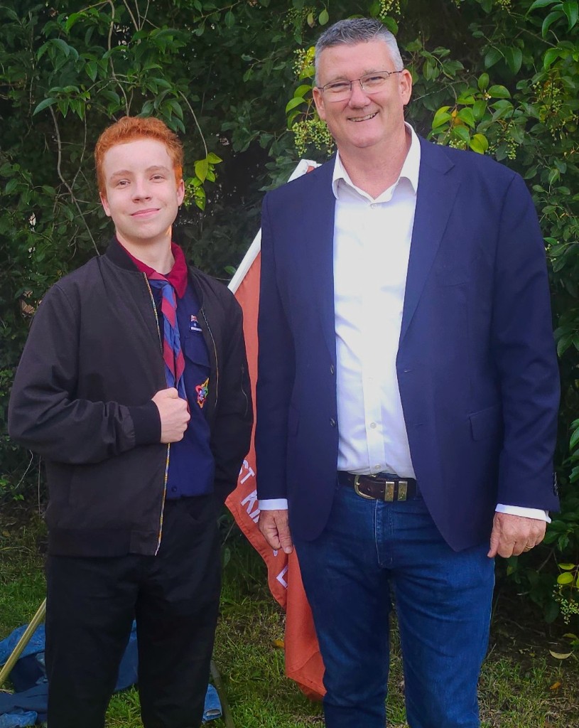 Eli Searle in scout neckerchief and jacket standing outdoors with Mark Taylor in a navy blazer and jeans — greenery behind